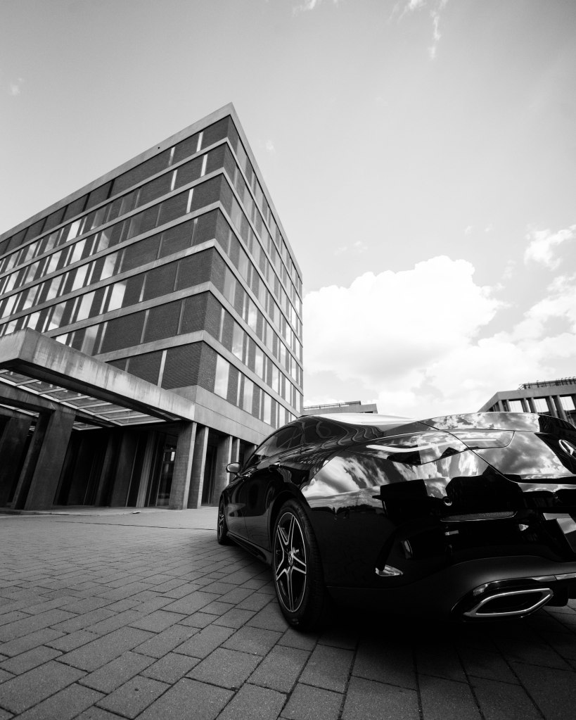 A black car parked on a paved area with a modern building in the background, captured in a black and white aesthetic.