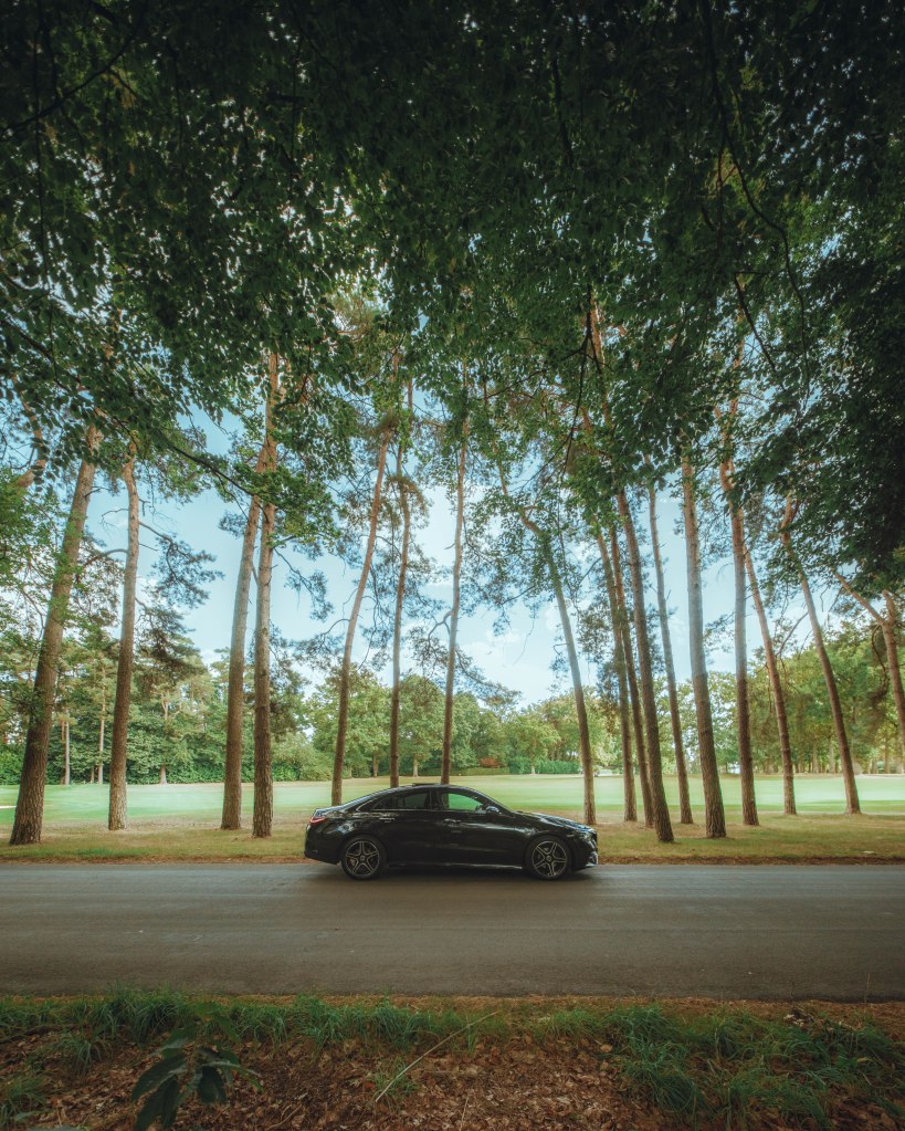 A black car parked on the side of a road lined with tall trees, with a grassy area in the background.
