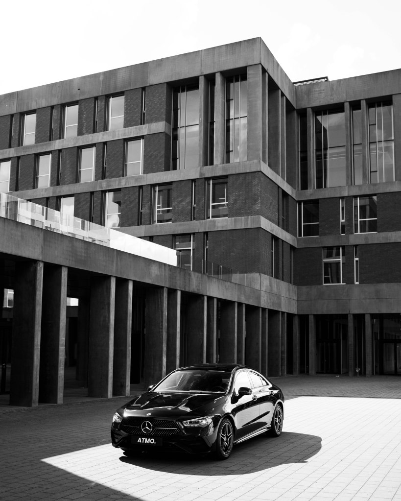 A black Mercedes-Benz parked in front of a modern building with large windows, captured in black and white.